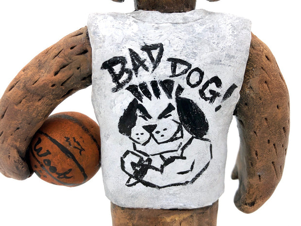 Sculpture of a fluffy tan dog with shaggy hair over its eyes. It stands like a human and wears a white tank top, a green backwards cap and holds a basketball under its arm.