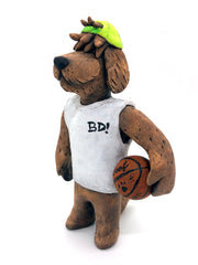 Sculpture of a fluffy tan dog with shaggy hair over its eyes. It stands like a human and wears a white tank top, a green backwards cap and holds a basketball under its arm.