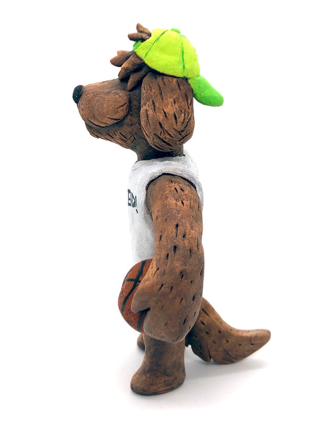 Sculpture of a fluffy tan dog with shaggy hair over its eyes. It stands like a human and wears a white tank top, a green backwards cap and holds a basketball under its arm.