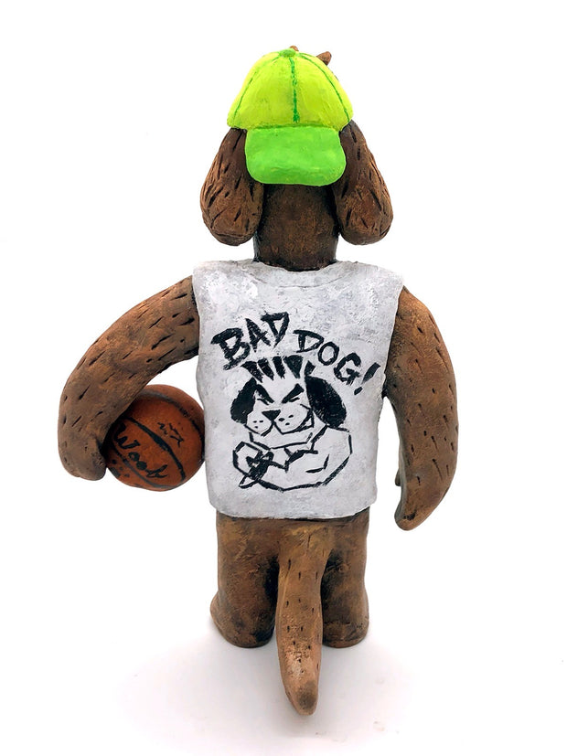 Sculpture of a fluffy tan dog with shaggy hair over its eyes. It stands like a human and wears a white tank top, a green backwards cap and holds a basketball under its arm.