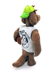 Sculpture of a fluffy tan dog with shaggy hair over its eyes. It stands like a human and wears a white tank top, a green backwards cap and holds a basketball under its arm.