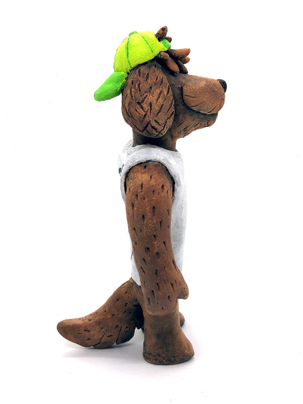 Sculpture of a fluffy tan dog with shaggy hair over its eyes. It stands like a human and wears a white tank top, a green backwards cap and holds a basketball under its arm.