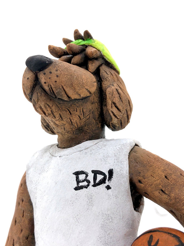 Sculpture of a fluffy tan dog with shaggy hair over its eyes. It stands like a human and wears a white tank top, a green backwards cap and holds a basketball under its arm.