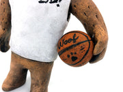 Sculpture of a fluffy tan dog with shaggy hair over its eyes. It stands like a human and wears a white tank top, a green backwards cap and holds a basketball under its arm.