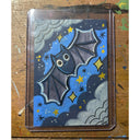 Artwork of a cartoon bat flying against a blue night sky with stars and clouds, photographed from above on a worn wooden surface.