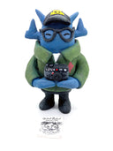Clay sculpture of a blue robot wearing glasses with a black baseball cap, green army jacket and holding a camera in both hands and a magazine in between its arm.