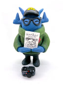 Clay sculpture of a blue robot wearing glasses with a black baseball cap, green army jacket and holding a camera in both hands and a magazine in between its arm.