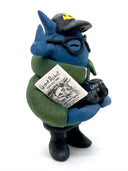 Clay sculpture of a blue robot wearing glasses with a black baseball cap, green army jacket and holding a camera in both hands and a magazine in between its arm.
