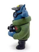 Clay sculpture of a blue robot wearing glasses with a black baseball cap, green army jacket and holding a camera in both hands and a magazine in between its arm.