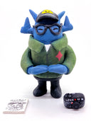 Clay sculpture of a blue robot wearing glasses with a black baseball cap, green army jacket and holding a camera in both hands and a magazine in between its arm.