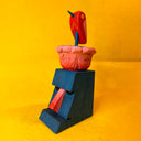 Colorful wooden sculpture of a tiki head with a red bird and pink flower atop its head, on a yellow background