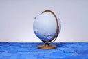 A globe, painting entirely light blue with a closed eye, calm facial expression. Small white wooden flowers are attached at opposite corners of the globe.