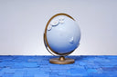 A globe, painting entirely light blue with a closed eye, calm facial expression. Small white wooden flowers are attached at opposite corners of the globe.