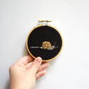 Small embroidery of a snail, with a trail of slime behind it. On a black background in an embroidery hoop.