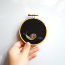 Embroidery of a snail with a tiny trail of slime behind it, on a black background in a wooden embroidery hoop.