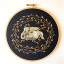 Embroidery on black fabric in a circular embroidery hoop of an upside down gray teapot, surrounded by fall colored leaves attached to vines.