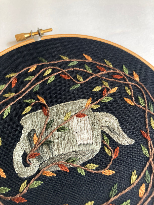 Embroidery on black fabric in a circular embroidery hoop of an upside down gray teapot, surrounded by fall colored leaves attached to vines.