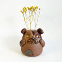Ceramic sculpture of a cute, brown bulldog with its paws on its cheek and a rounded bottom without legs. It has flowers sticking out of its head, like a vase.
