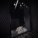 A black arched mirror with a thin metallic frame, set against a dark quilted black wall. The mirror reflects a black marble floor and wall, decorated with white star decals near the top and a white line-art illustration of a 2 headed calf near the bottom.