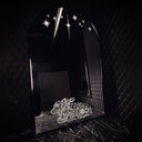 A black arched mirror with a thin metallic frame, set against a dark quilted black wall. The mirror reflects a black marble floor and wall, decorated with white star decals near the top and a white line-art illustration of a 2 headed calf near the bottom.