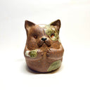 Ceramic sculpture of a cute, brown cat with its paws on its cheek and a rounded bottom without legs. It has green textured spots.