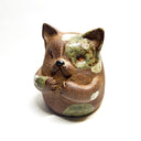 Ceramic sculpture of a cute, brown cat with its paws on its cheek and a rounded bottom without legs. It has green textured spots.
