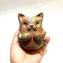 Ceramic sculpture of a cute, brown cat with its paws on its cheek and a rounded bottom without legs. It has green textured spots.