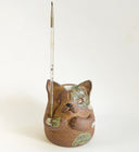 Ceramic sculpture of a cute, brown cat with its paws on its cheek and a rounded bottom without legs. It has green textured spots.