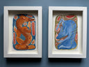 Two framed abstract paintings hanging side by side on a dark blue-gray wall. The left print shows a stylized orange creature on a blue background, and the right print shows a stylized blue creature on a tan background.