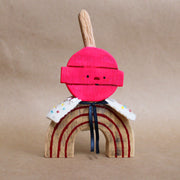 Wooden sculpture of a pink round lollipop, with its stick up in the air. Its round head is attached to an arch shaped body with no limbs. It wears a white cape with colorful sparkles.