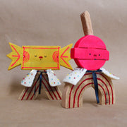 Wooden sculpture of a pink round lollipop, with its stick up in the air. Its round head is attached to an arch shaped body with no limbs. It wears a white cape with colorful sparkles.