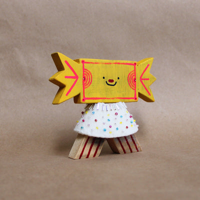 Wooden sculpture of a yellow wrapped piece of candy with a painted on face. Its head is attached to a triangular shaped body with no limbs. It wears a white cape with colorful sparkles.