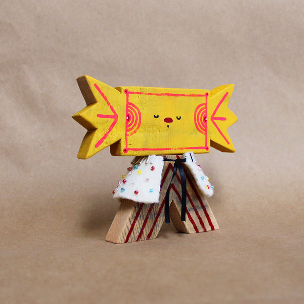 Wooden sculpture of a yellow wrapped piece of candy with a painted on face. Its head is attached to a triangular shaped body with no limbs. It wears a white cape with colorful sparkles.