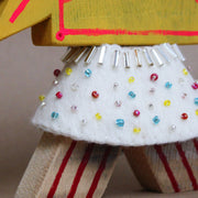 Wooden sculpture of a yellow wrapped piece of candy with a painted on face. Its head is attached to a triangular shaped body with no limbs. It wears a white cape with colorful sparkles.