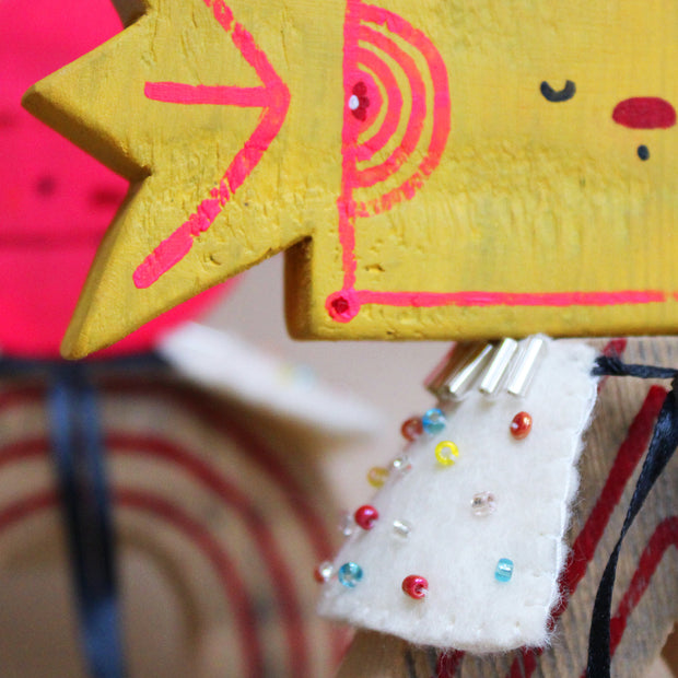 Wooden sculpture of a yellow wrapped piece of candy with a painted on face. Its head is attached to a triangular shaped body with no limbs. It wears a white cape with colorful sparkles.