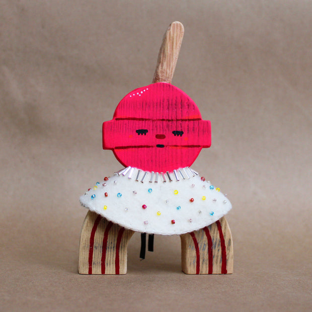 Wooden sculpture of a pink round lollipop, with its stick up in the air. Its round head is attached to an arch shaped body with no limbs. It wears a white cape with colorful sparkles.