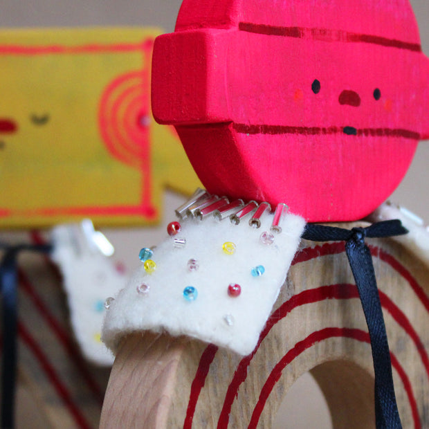 Wooden sculpture of a pink round lollipop, with its stick up in the air. Its round head is attached to an arch shaped body with no limbs. It wears a white cape with colorful sparkles.