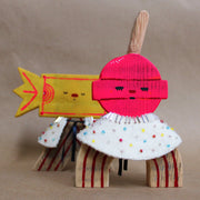 Wooden sculpture of a pink round lollipop, with its stick up in the air. Its round head is attached to an arch shaped body with no limbs. It wears a white cape with colorful sparkles. 