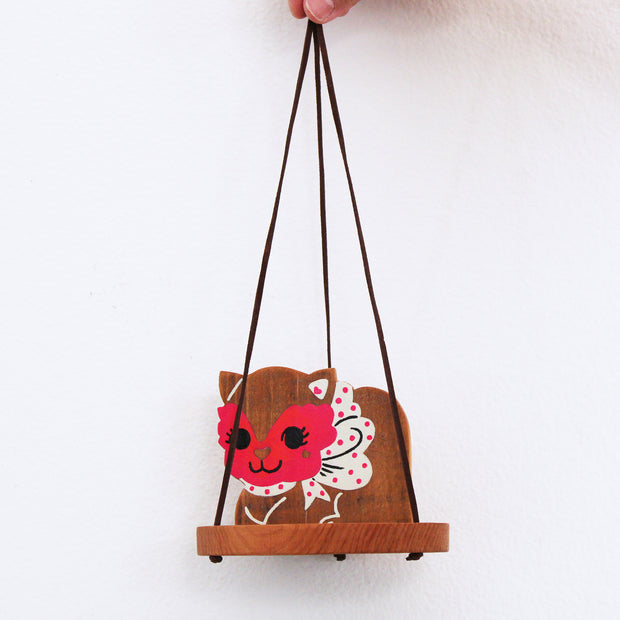 Wooden sculpture of a cat with a red masquerade style mask on, a white and pink polka dot bow and a smile. It sits on a round tray and hangs from 3 suede strings.