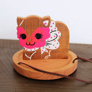 Wooden sculpture of a cat with a red masquerade style mask on, a white and pink polka dot bow and a smile. It sits on a round tray and hangs from 3 suede strings.