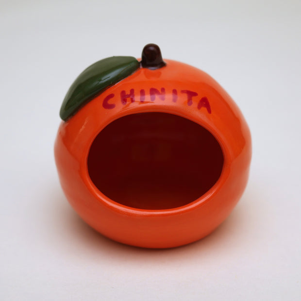 Small ceramic sculpture of an orange or tangerine, with a very short stem and a green leaf. It has decorative red writing on the front and back and a hole carved out of the front.