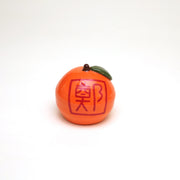 Small ceramic sculpture of an orange or tangerine, with a very short stem and a green leaf. It has decorative red writing on the front and back and a hole carved out of the front.