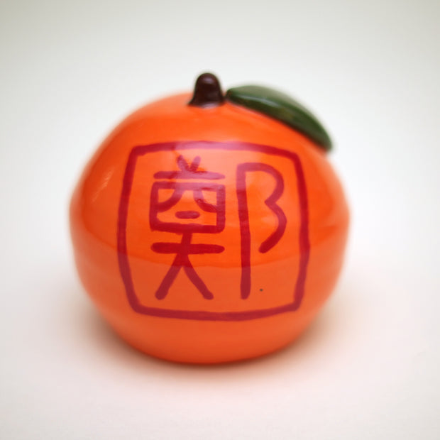 Small ceramic sculpture of an orange or tangerine, with a very short stem and a green leaf. It has decorative red writing on the front and back and a hole carved out of the front.