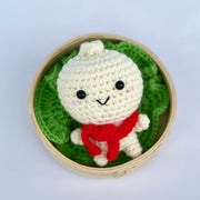 A crocheted bao dumpling, with a smiling cute chibi face with pink cheeks. It wears a red knit scarf and sits inside of a bao steamer atop of a knit lettuce leaf.