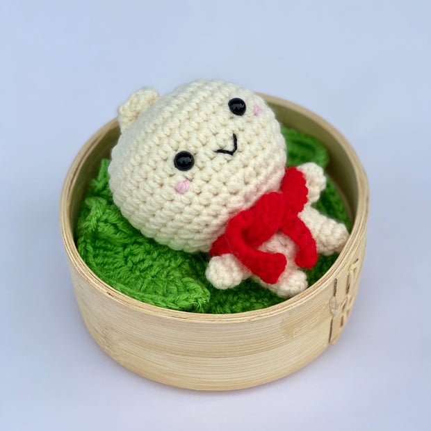 A crocheted bao dumpling, with a smiling cute chibi face with pink cheeks. It wears a red knit scarf and sits inside of a bao steamer atop of a knit lettuce leaf.