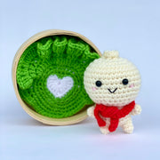 A crocheted bao dumpling, with a smiling cute chibi face with pink cheeks. It wears a red knit scarf and stands outside of a bao steamer atop of a knit lettuce leaf with a white heart in the center.