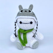 Crocheted version of Baymax from Big Hero 6, a white round edged robot with a kind face. It wears a green knit scarf and a crocheted beanie atop its head that resembles Totoro.