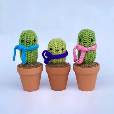 3 crocheted cacti in real clay pots, with crocheted soil and crocheted scarfs. Scarf colors are blue, purple or pink.