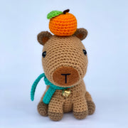 Crochet sculpture of a sitting capybara with an oversize head and smaller body. Atop its head is an orange citrus fruit and around its neck is a teal scarf and a metal bell. 