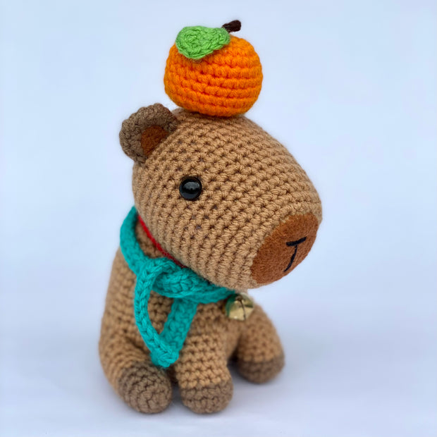 Crochet sculpture of a sitting capybara with an oversize head and smaller body. Atop its head is an orange citrus fruit and around its neck is a teal scarf and a metal bell.